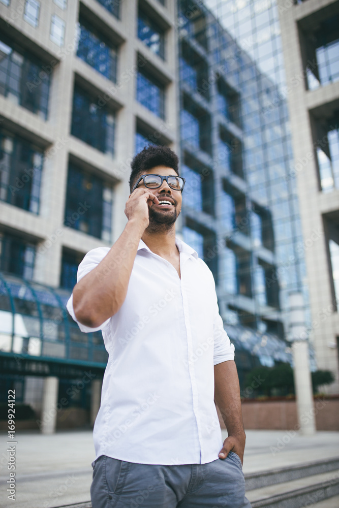 Young handsome Afro American man standing in front of huge modern business building smiling and talking on cell phone Young handsome Afro American man standing in front of huge modern business building smiling and talking on cell phone