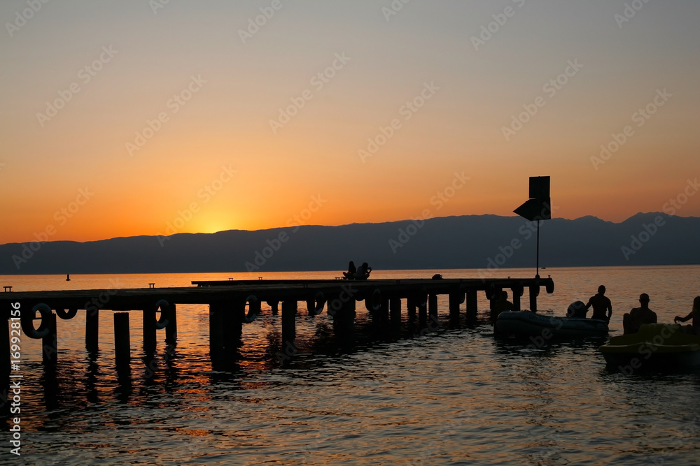Silhouettes of boat dock and young people on sunset; summer landscape background.