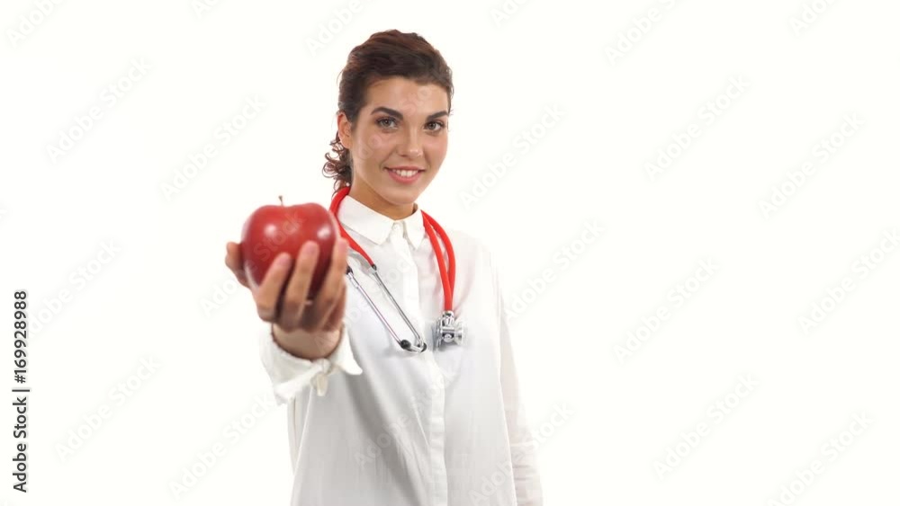 Young friendly female nutritionist showing a red apple, advising healthy diet. Portrait of young professional with stethoscope and lab coat isolated on white background Young friendly female nutritionist showing a red apple, advising healthy diet. Portrait of young professional with stethoscope and lab coat isolated on white background