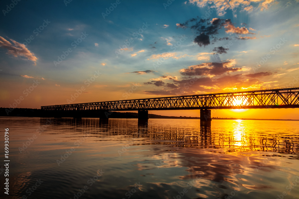 Fototapeta premium Stunning sunset colors over river with bridge in a summer evening. Dramatic clouds. Toned.