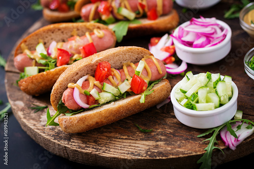 Hot dog with cucumber, tomato and red onion on wooden background.