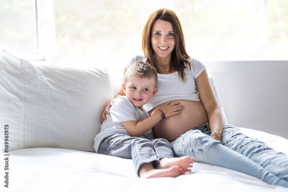 Beautiful pregnant young mother, sitting in bed with her older child Beautiful pregnant young mother, sitting in bed with her older child