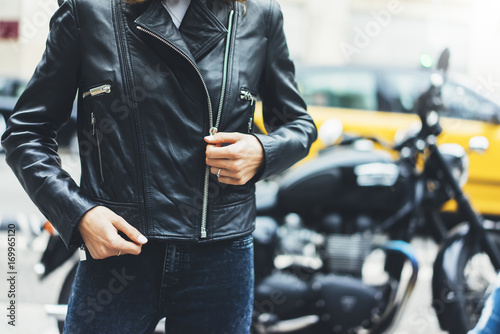 Girl unfastens black leather jacket on background motorcycle in sun flare city, hipster biker female hands closeup, motorbike street lifestyle, traveler planing bike route in summer holiday concept photo