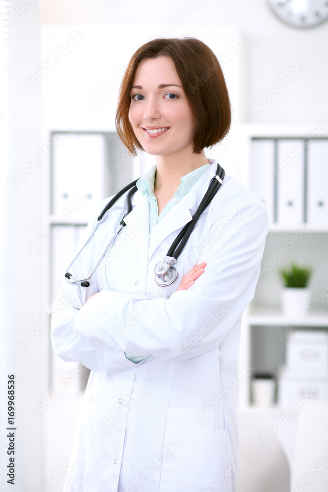 Young brunette female doctor standing with arms crossed and smiling at hospital Young brunette female doctor standing with arms crossed and smiling at hospital