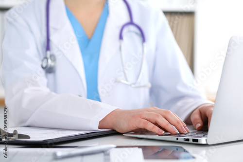 Close up of unknown female doctor typing on laptop computer while sitting at the table