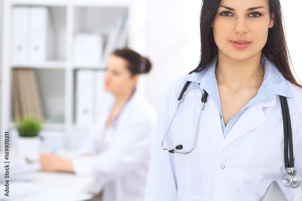 Brunette female doctor on the background of colleagues talking to each other in hospital. Physician is ready to help patients. Medicine and health care concept Brunette female doctor on the background of colleagues talking to each other in hospital. Physician is ready to help patients. Medicine and health care concept