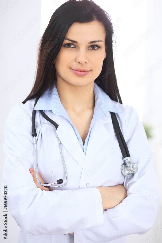 Brunette female doctor on the background of colleagues talking to each other in hospital. Physician is ready to help patients. Medicine and health care concept