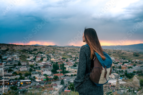 Fototapeta Naklejka Na Ścianę i Meble -  Young woman tourist from a high point looking at the sunset over the city of Goreme in Turkey and dreaming. Cappadocia. Tourism, rest, vacation.