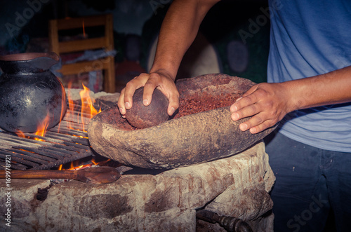 People grinding a cacao beans over a rock, next to a wood stove