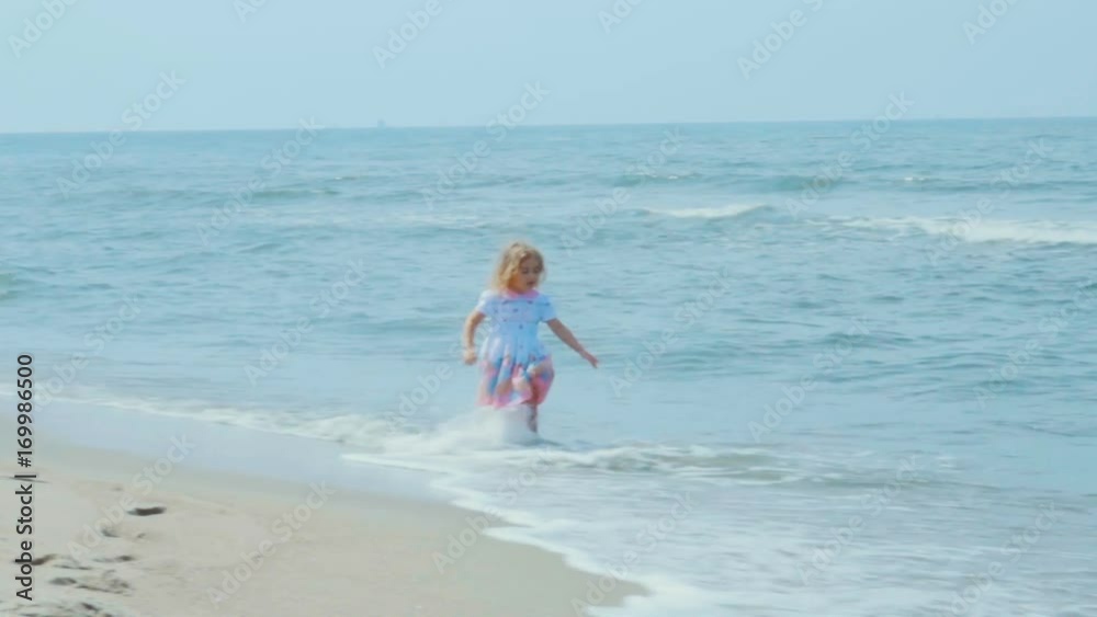 Adorable little girl in a dress runs jumping on the beach. Child enjoys playing on the seashore running on the water with pleasure. splashing in sea waves. Adorable little girl in a dress runs jumping on the beach. Child enjoys playing on the seashore running on the water with pleasure. splashing in sea waves.