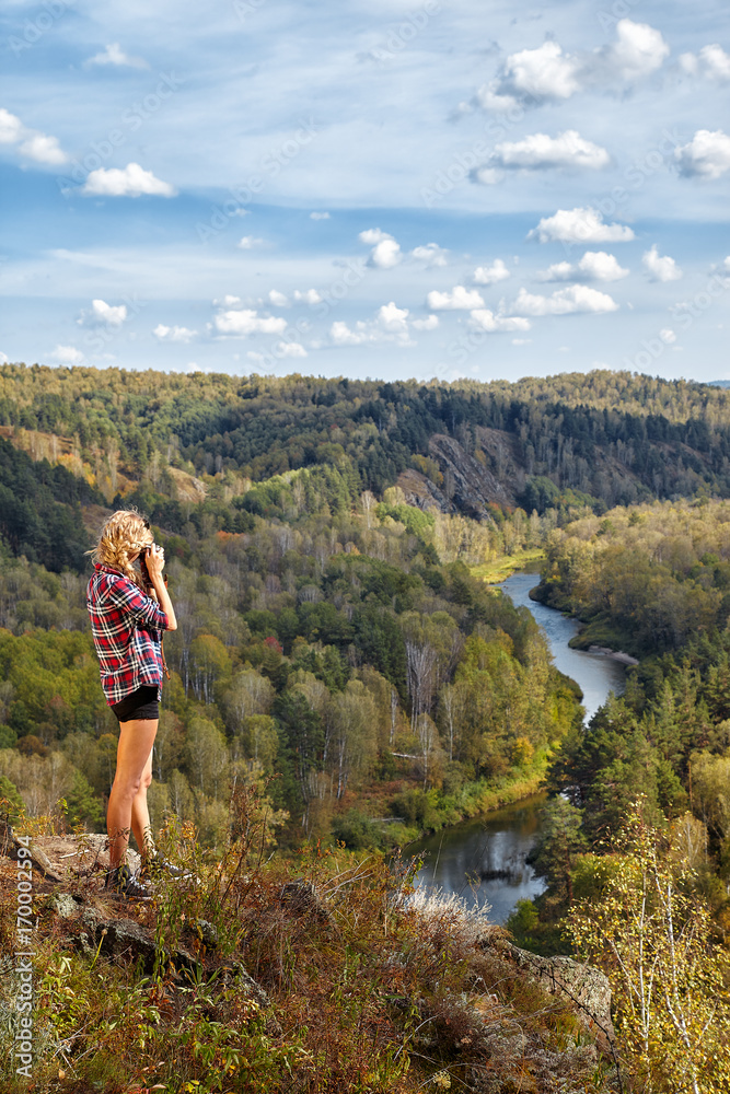 Young blonde woman tourist   on a cliff taking pictures of the autumn landscape with the river Berd on old film camera