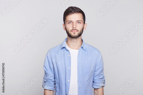 Portrait of Caucasian handsome brunette  guy with blue eyes, trendy hairdo and beard looking seriously at camera while posing against white studio wall. Grave businessman in shirt thinking about work.