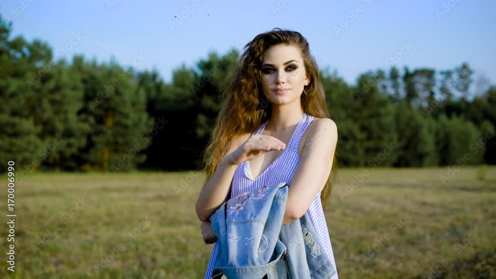 A young woman in a summer sarafan pensively stands in the nature on a summer day, a lady dresses a denim jacket on her shoulder A young woman in a summer sarafan pensively stands in the nature on a summer day, a lady dresses a denim jacket on her shoulder