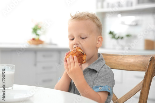 Cute little boy eating big muffin in kitchen