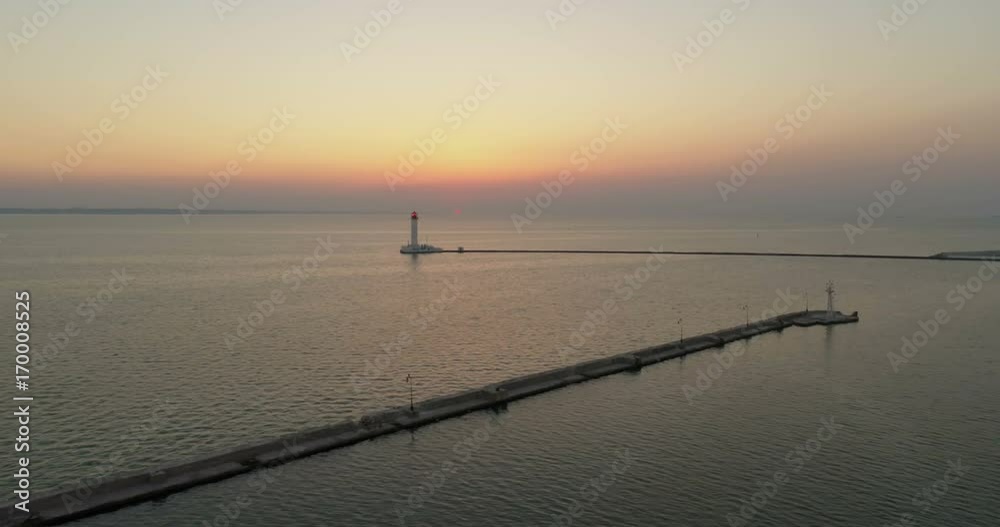 Drone flies sideways over the tranquil waters of Odessa Port with the Odessa Lighthouse behind. The sun is rising in the distance Drone flies sideways over the tranquil waters of Odessa Port with the Odessa Lighthouse behind. The sun is rising in the distance
