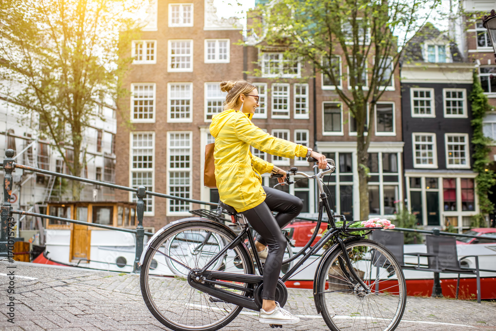 Naklejka premium Młoda kobieta w żółtym płaszczu przeciwdeszczowym z torbą i kwiatami, jazda na rowerze w mieście Amsterdam