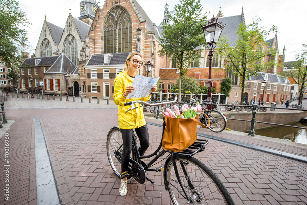 Naklejka premium Portret turystki kobieta w żółtym płaszczu stojącej z mapą i rowerem ze starym kościołem na tle w Amsterdamie