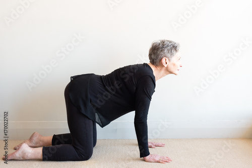 Side view of older woman with short grey hair and black clothing in yoga cat stretch posture