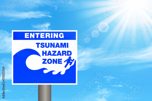 Tsunami hazard zone warning sign on blue sky and sun light white clouds background.