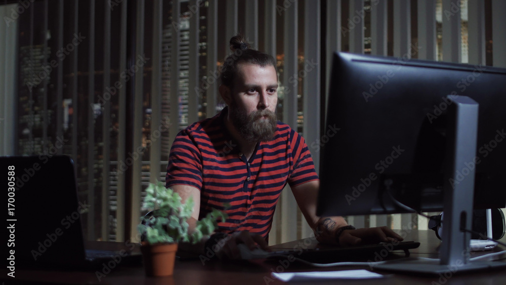 Handsome bearded man sitting at computer in office and working in the evening. Handsome bearded man sitting at computer in office and working in the evening.