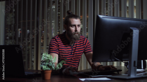 Handsome bearded man sitting at computer in office and working in the evening.