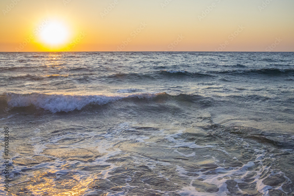 Panorama del sole che tramonta a mare. L'acqua è mossa e le onde a contatto con la sabbia formano la schiuma. Il mare è agitato e il cielo è arancione per via del crepuscolo. Panorama del sole che tramonta a mare. L'acqua è mossa e le onde a contatto con la sabbia formano la schiuma. Il mare è agitato e il cielo è arancione per via del crepuscolo.