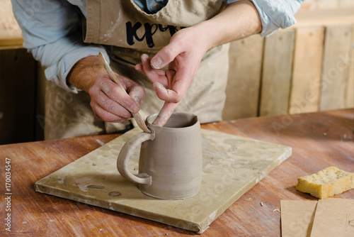 potter, workshop, ceramics art concept - ceramist holding cup and carefully examines the product, young male master dressed in a stained clay apron, craftsman's hands holding unbaked jug