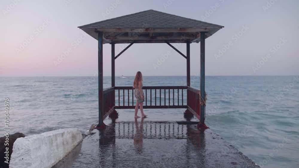 Young beautiful woman walking on pier in midi dress at sunset. Attractive blond girl standing near storm waves hitting the pier. Waves splashes in slow motion. Freedom,solitude concept. Young beautiful woman walking on pier in midi dress at sunset. Attractive blond girl standing near storm waves hitting the pier. Waves splashes in slow motion. Freedom,solitude concept.