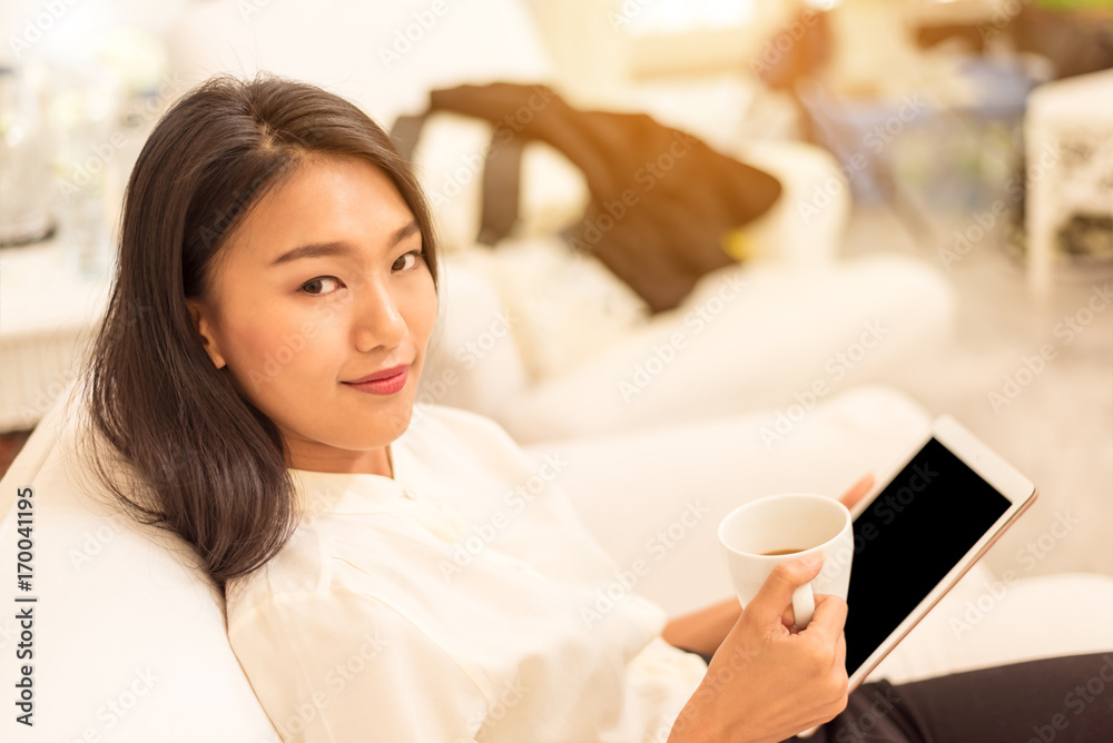 Young asian businesswoman holding mug of coffee and tablet computer in her hands .She sitting on white sofa at home in morning time. Young asian businesswoman holding mug of coffee and tablet computer in her hands .She sitting on white sofa at home in morning time.