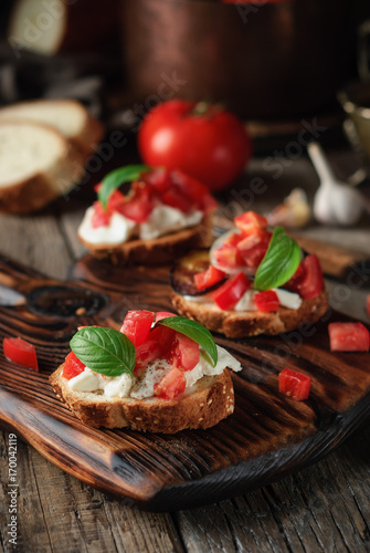 Homemade appetizer bruschetta with ciabatta, feta cheese, fresh tomatoes and green basil leaves on a wooden cutting board and ingredients on a gark grey wooden background. Close up photo