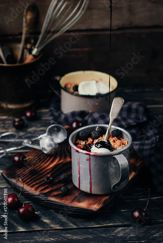 Crumbles of oatmeal, cherries, blackberries and black currants with vanilas ice cream and chocolate topping in vintage metal mugs on a black wooden background. Close up. Toned picture photo