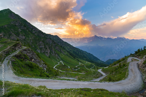 Dirt mountain road leading to high mountain pass in Italy (Colle delle Finestre). Expasive view at sunset, colorful dramatic sky, adventures in summer time, Italian Alps.