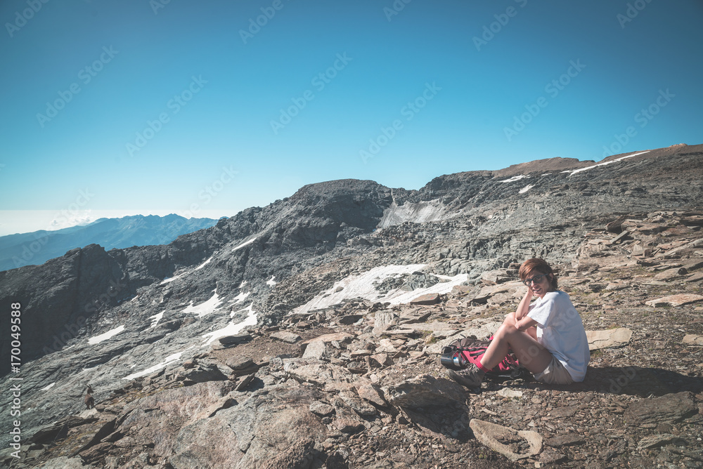 One person sitting on rocky terrain and watching a colorful sunrise high up in the Alps. Wide angle view from above with glowing mountain peaks in the background. Toned image.