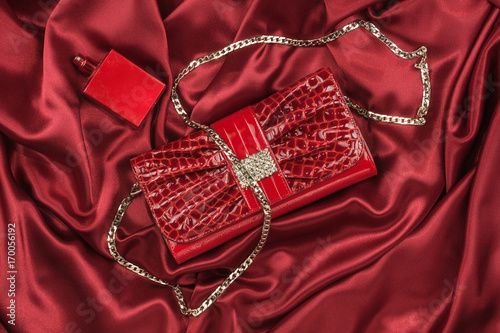 Bag of lacquered leather and red perfume lying on red silk. Handbag for women and bottle of scent, top view.