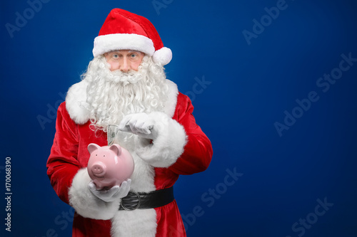 Santa Claus putting dollar into piggy bang on color background