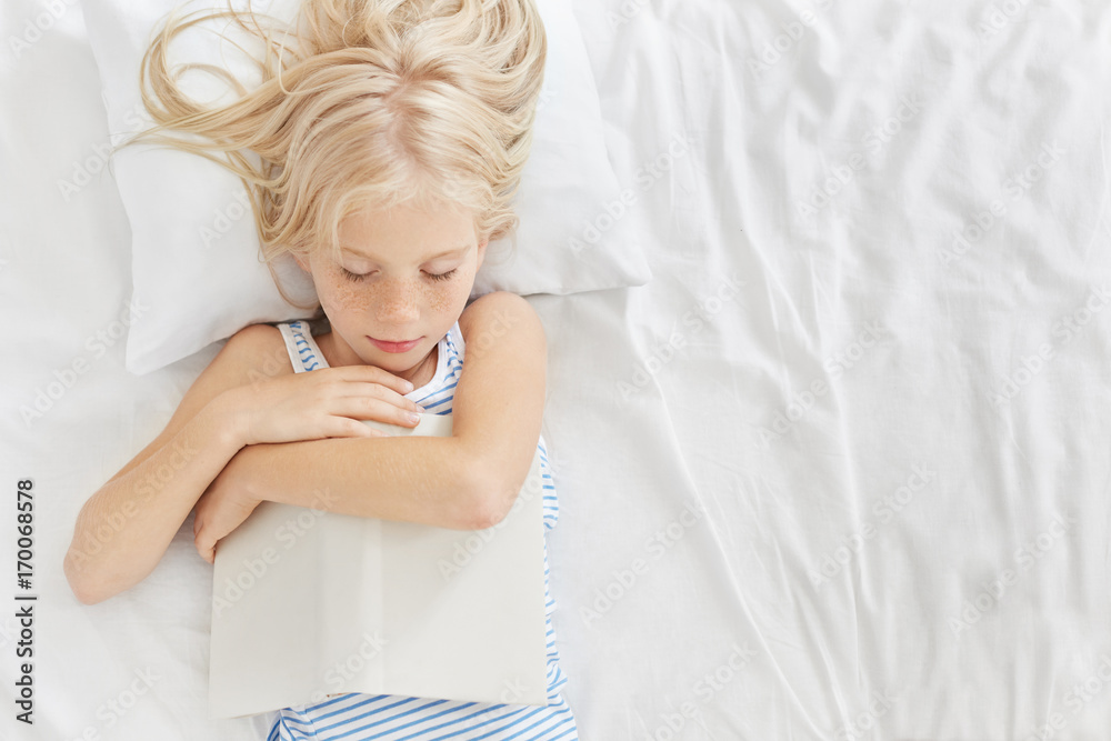 Sleeping girl with long blonde hair, keeping book in hands, falling asleep after reading fantasy or fairy tales, having pleasant dreams. Kid resting in comfortable room after active play with friends Sleeping girl with long blonde hair, keeping book in hands, falling asleep after reading fantasy or fairy tales, having pleasant dreams. Kid resting in comfortable room after active play with friends