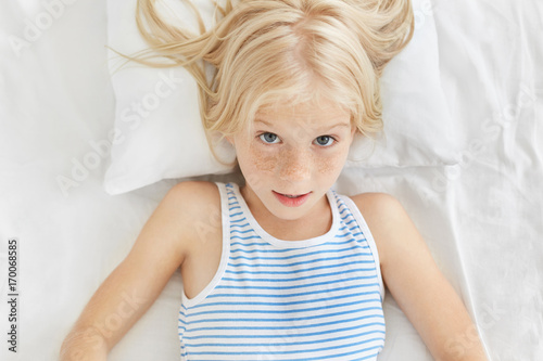 Portrait of cute blonde girl wearing sailor t-shirt, looking surprisedly into camera, awaking in morning hearing loud alarm cloak. Adorable girl feeling comfort while resting in bed in her room photo