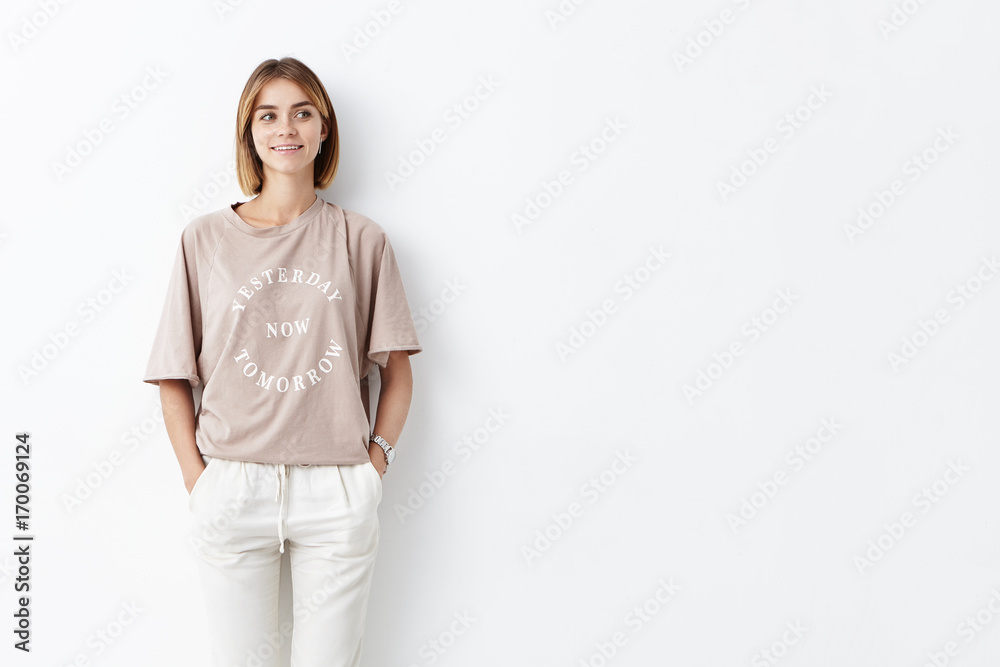 Attractive young female model with bobbed hair, wearing stylish clothes, keeping her hands in pockets, posing against white background with copy space for your promotional text or advertisment Attractive young female model with bobbed hair, wearing stylish clothes, keeping her hands in pockets, posing against white background with copy space for your promotional text or advertisment