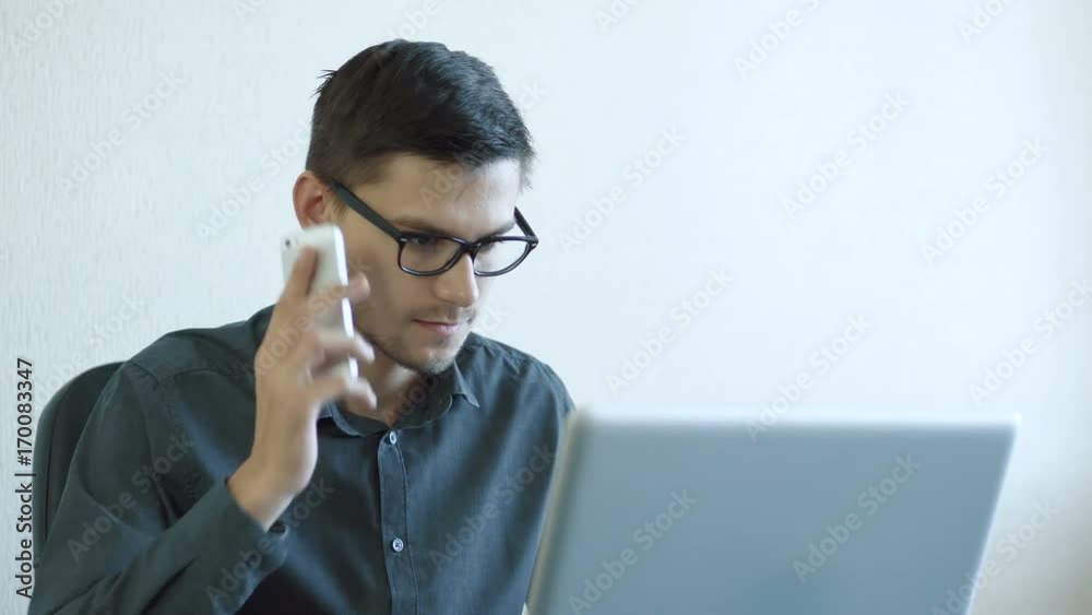 Portrait of a young man wearing glasses sitting in his office in front of a monitor - working on a computer and talking on the phone. People stock footage slider shot. Portrait of a young man wearing glasses sitting in his office in front of a monitor - working on a computer and talking on the phone. People stock footage slider shot.