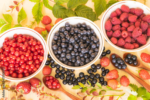 Fresh and colorful berries assortment with green leaves on the light wooden background.