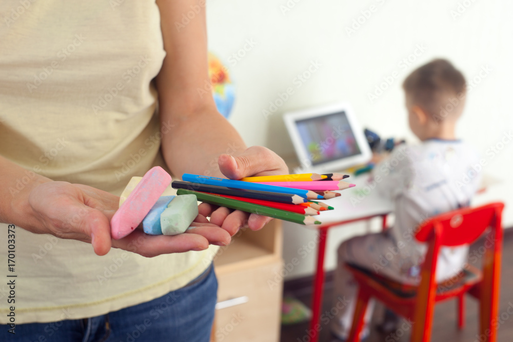 woman offers pencils and crayons for drawing on the background of a drawing boy, selective focus