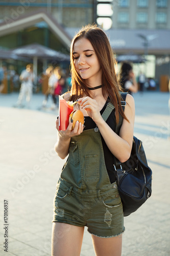 A young girl with a cheeseburger and fries in her hand walks around the city square