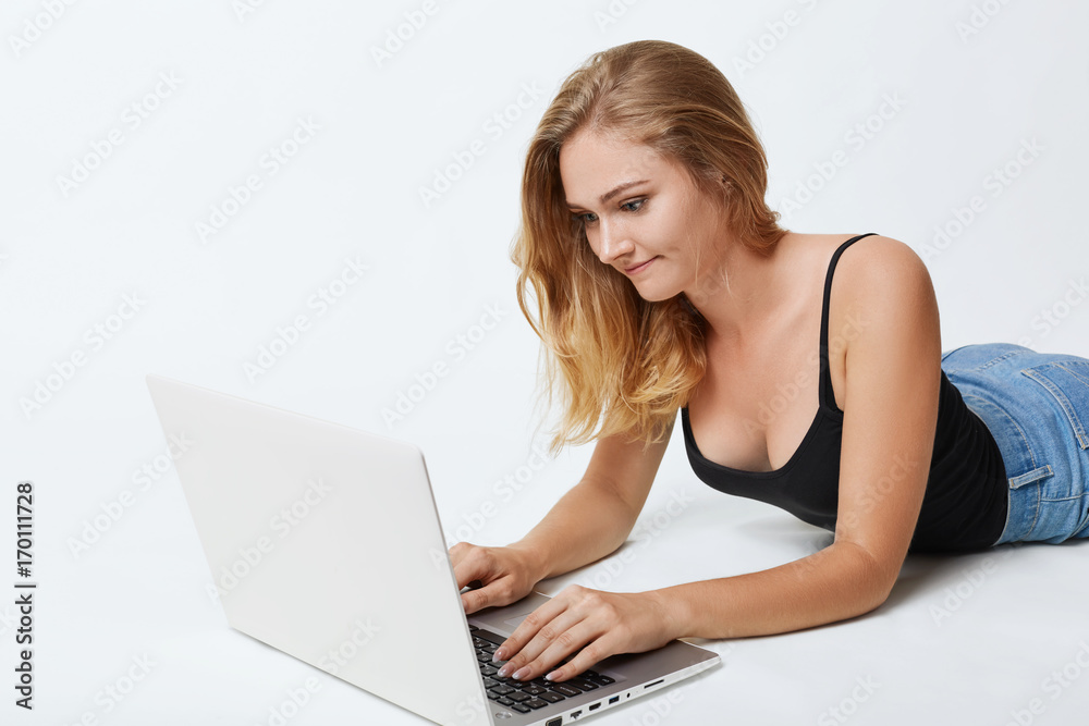 Restful female with long blonde hair, lying on white floor in front of opened laptop, messaging with friends in social networks, having concentrated and focused look into screen. Communication concept Restful female with long blonde hair, lying on white floor in front of opened laptop, messaging with friends in social networks, having concentrated and focused look into screen. Communication concept