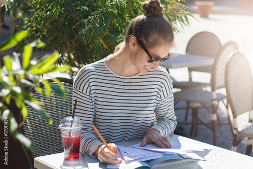 Closeup image of young female wearing trendy sunglasses in street cafe in summer  bending to papers and books lying on table filling important data and checking facts  looking happy and relaxed