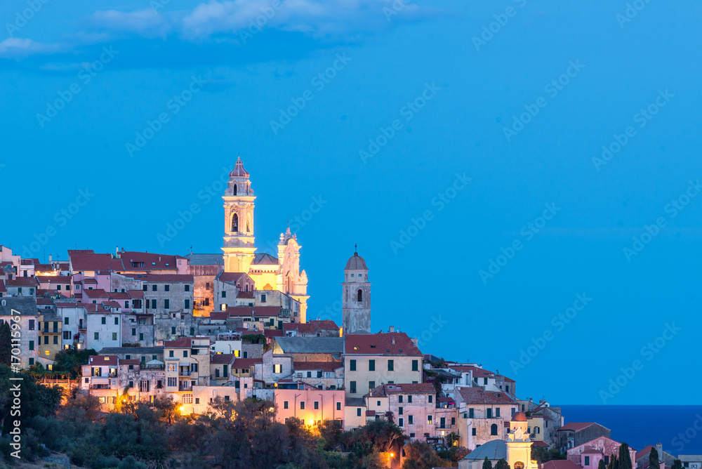 The old town of Cervo, Liguria, Italy, with the beautiful baroque church and tower bells arising from the colorful houses, illuminated at twilight.