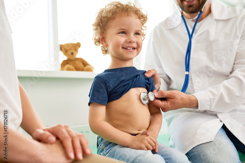 Portrait of adorable little boy visiting doctor, looking brave and smiling, holding  while pediatrician listening to heartbeat with stethoscope photo