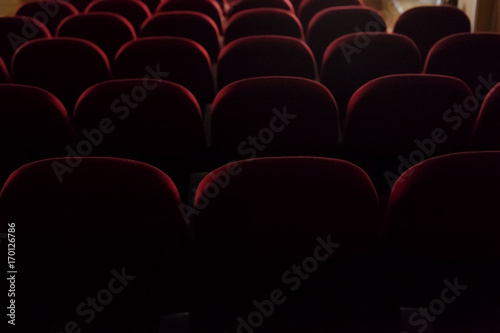Back view of many rows of red velvet seats in a cinema or theater stage. Semi-darkness.
