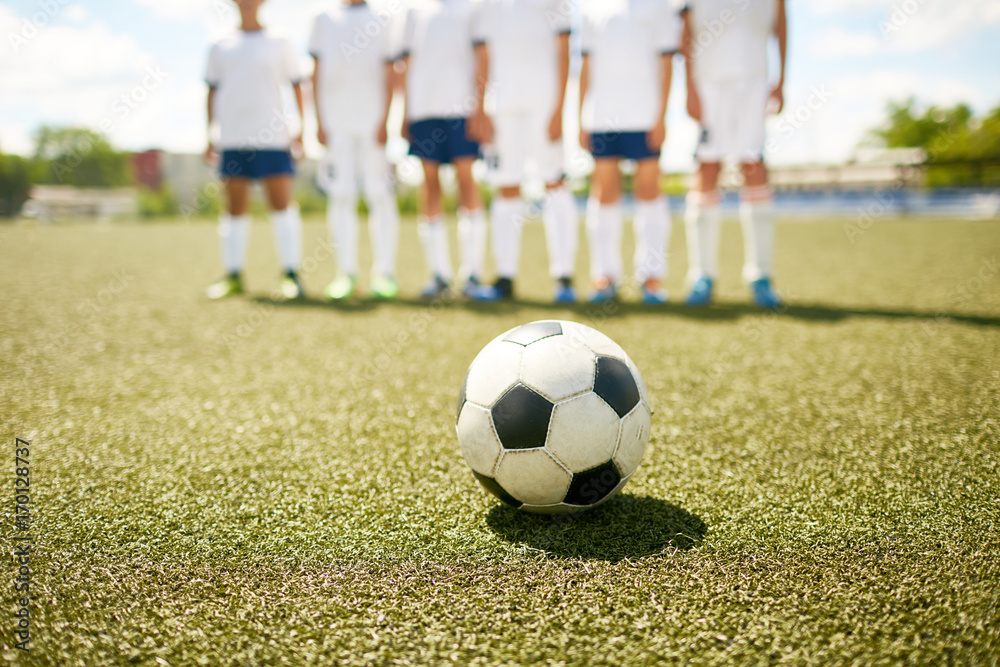 Fototapeta premium Closeup of football ball lying on field with blurred junior football team in background, preparing for game