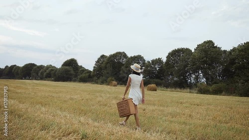 A young girl wearing a white dress with a straw hat and a suitcase. Straw and straw bales. Sunset photo