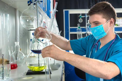 Young male scientist in uniform wearing a mask is holding test glass and filling a test tube with blue liquid